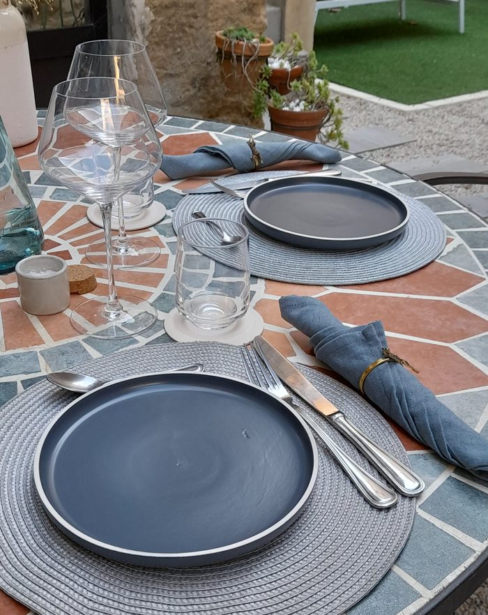 Ronds de table osier gris, assiettes bleu, couverts inox, verres à vins et eau, serviette bleu jean sur table ronde mosaïque orange et grise