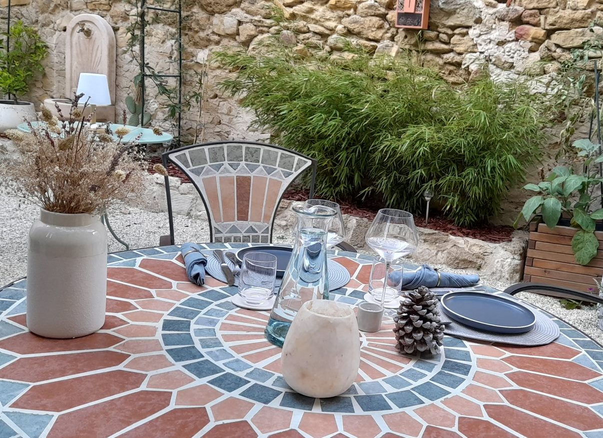 Table ronde en fer forgé avec une rosace en carrelage dans les teintes bleues, orangées, avec les assiettes, les verres et les couverts et un vase en grey blanc avec un bouquet de fleurs séchées. Une fontaine et des bambous en arrière plan